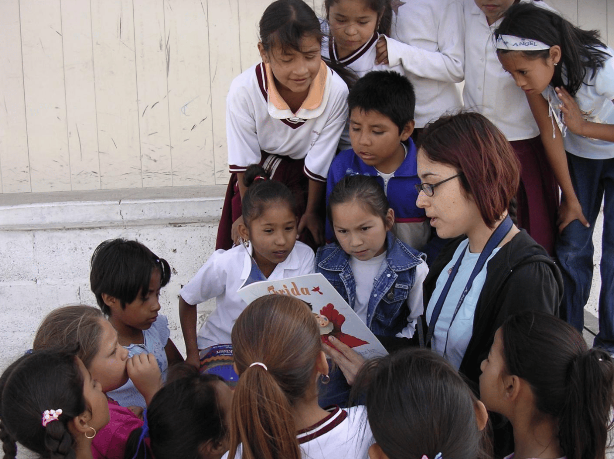 Adult woman reads a book titled “Frida” to a group of children outdoors.