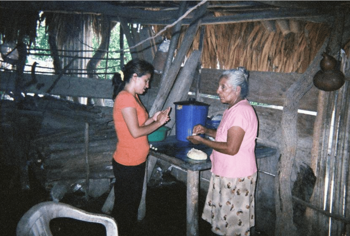 Two women stand beside a rough wooden table in a dim, thatched rural kitchen.